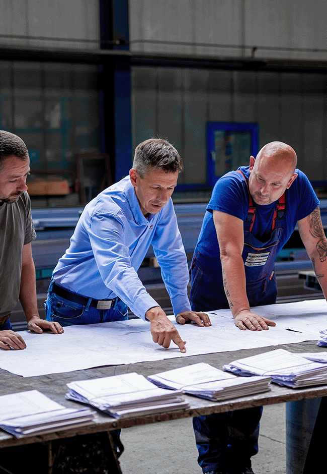 Projektbesprechung von drei Mitarbeitern über technische Pläne in der Werkhalle