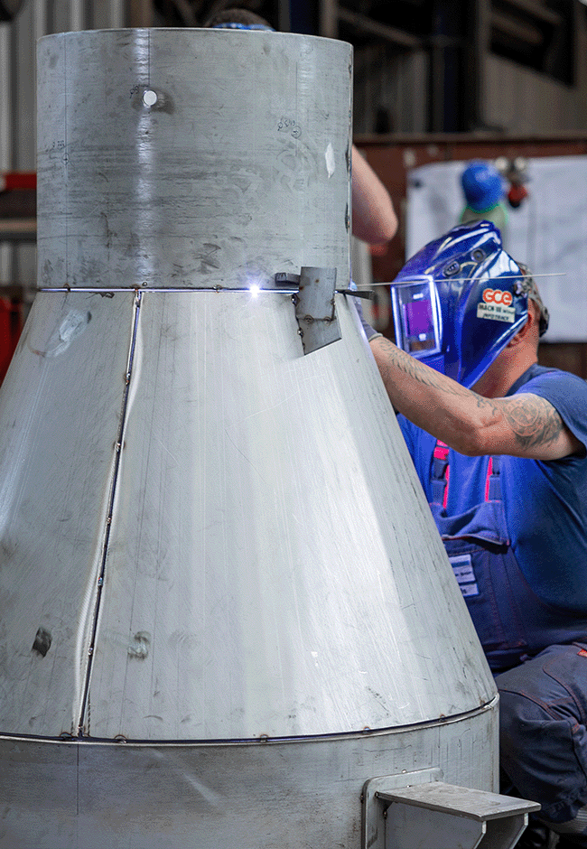 Schweißer bearbeitet große Metallkonstruktion mit Schutzhelm in der Werkhalle